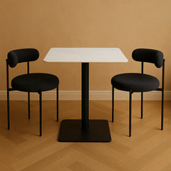 Two black chairs with cushioned seats and backrests are arranged around a white square table on a wooden floor.